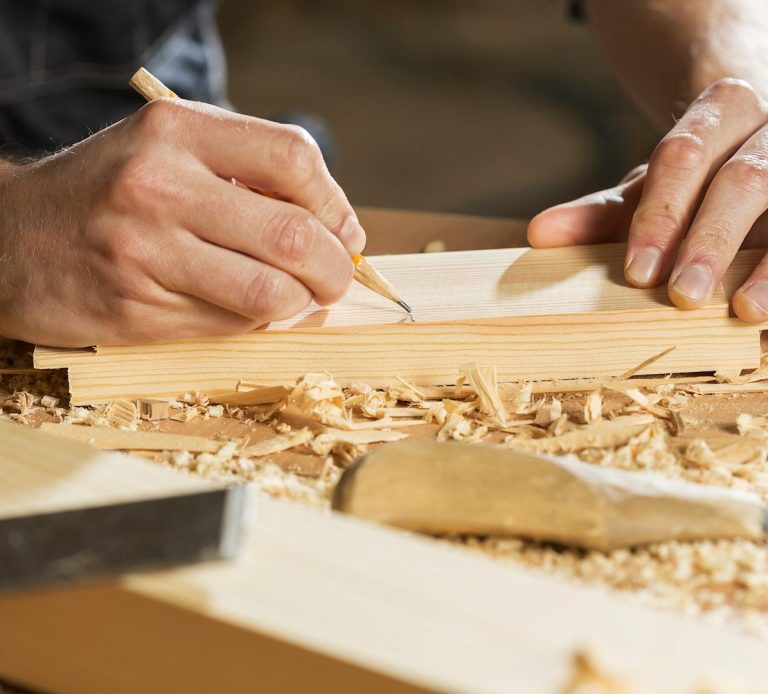 A carpenter's hands marking wood on a workbench with shavings scattered around.