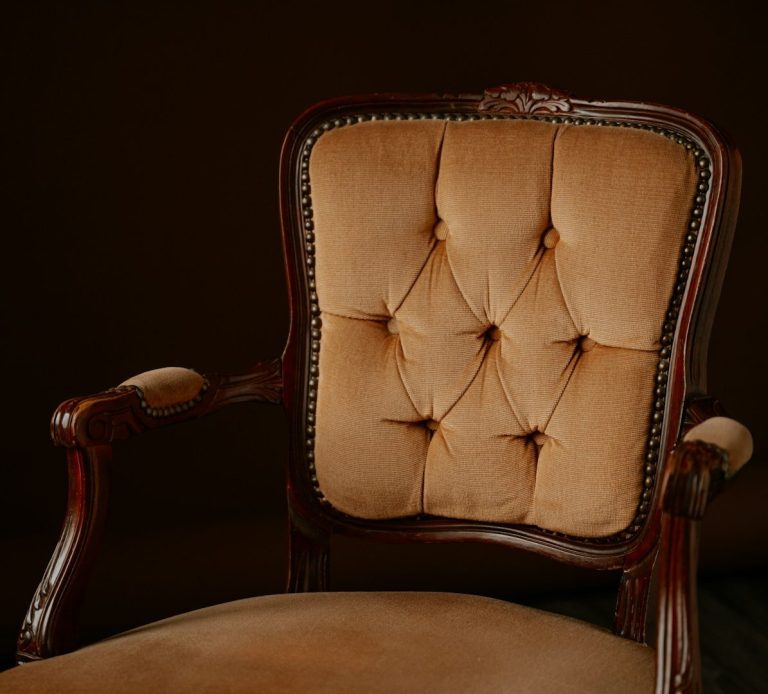 Antique wooden armchair with tufted, beige upholstery against a dark background.