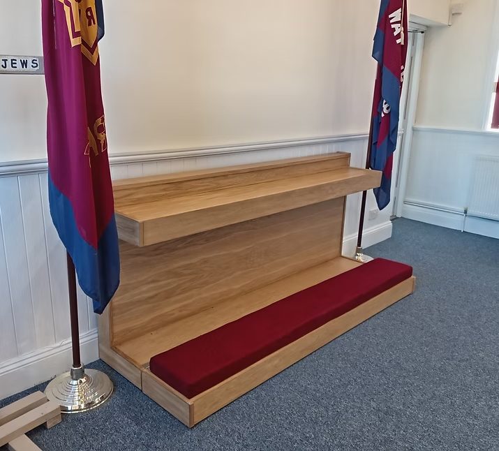 Wooden mercy seat with a red cushion, flanked by Salvation Army flags, in a light-coloured room.